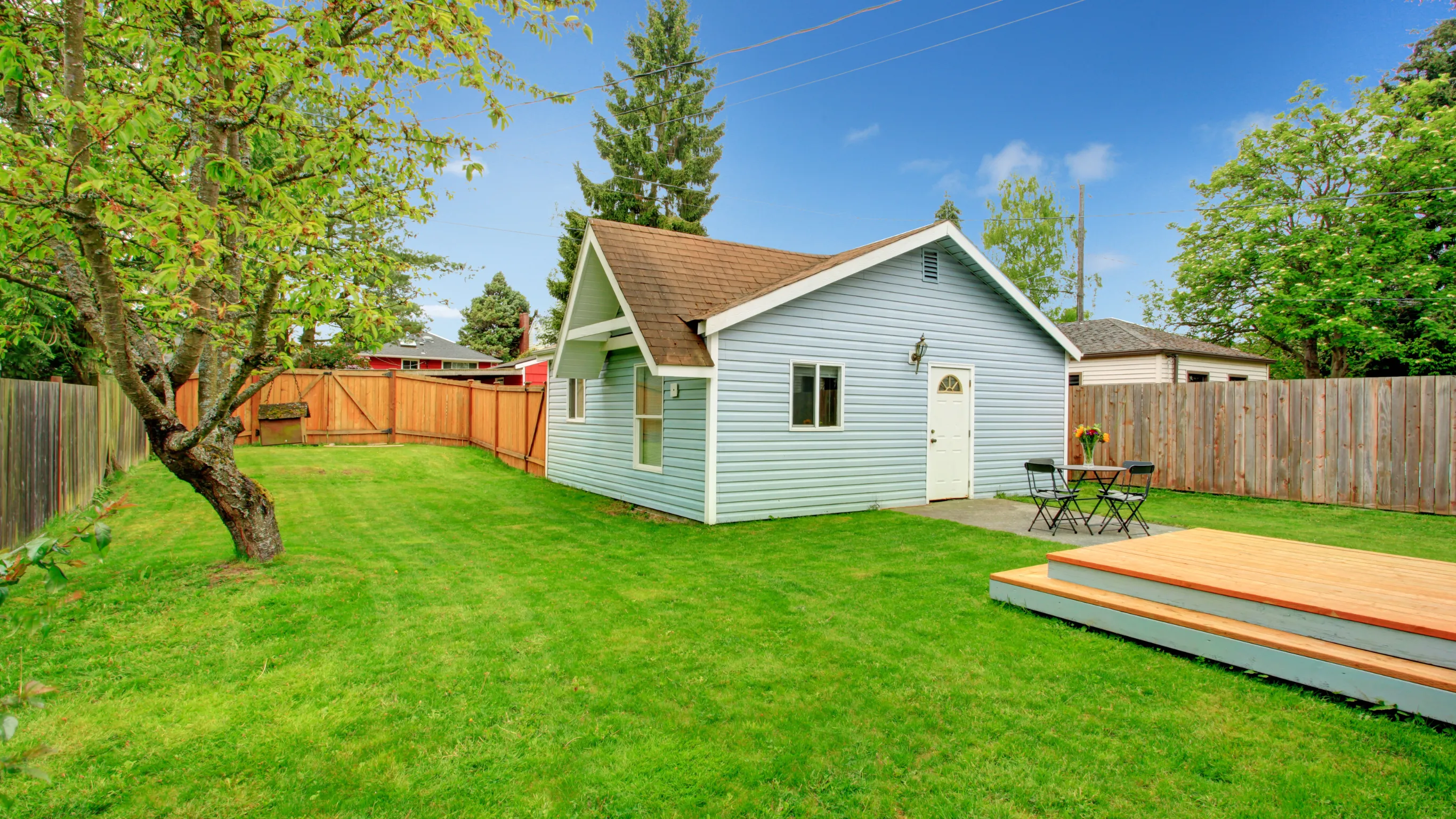 Backyard guest house project in Los Angeles showing a private cottage style guest unit with light blue siding, white trim, small patio door, fenced yard, green lawn, and outdoor deck